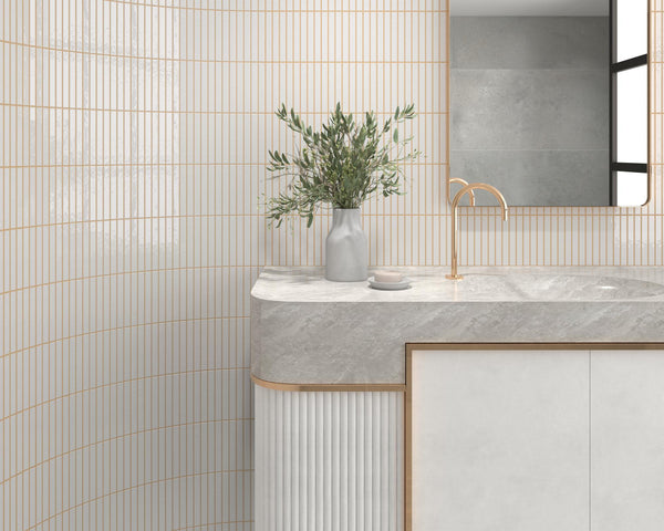 A modern bathroom with vertical white and gold tiles, a marble countertop, a gold faucet, a rectangular mirror, and a vase with green branches on the counter.