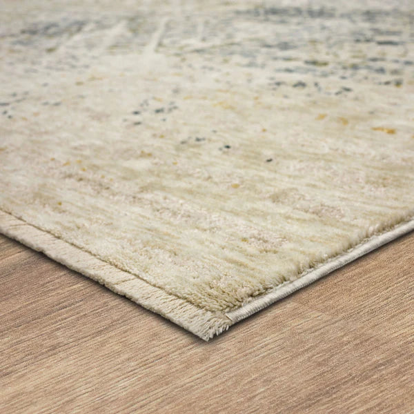 A close-up of the corner of a light-colored, textured area rug with subtle patterns, laying on a wooden floor.