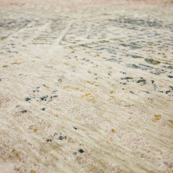 Close-up view of a textured area rug with a mix of beige, cream, blue, and subtle yellow tones in an abstract, faded pattern. The surface appears soft and slightly plush.