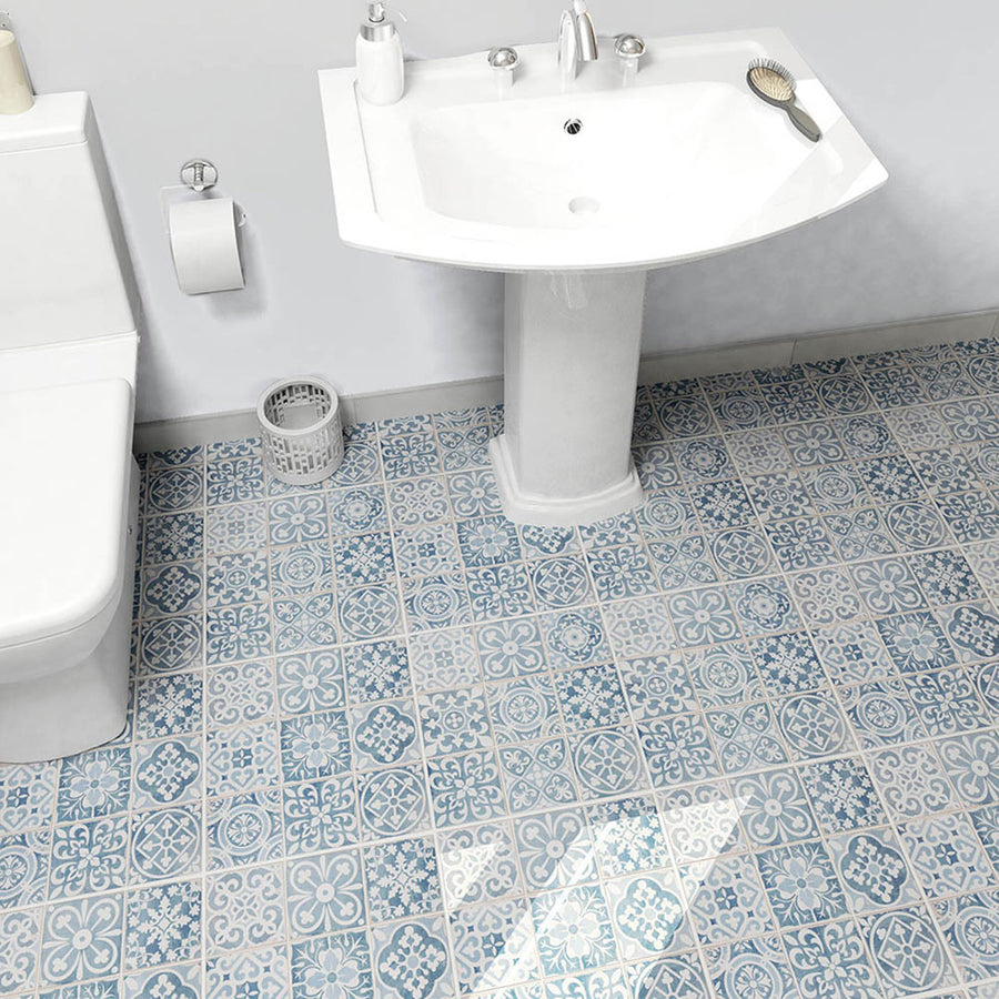 Blue and white patterned tile bathroom floor with a white pedestal sink, toilet, toilet paper holder, trash bin, soap dispenser, and a hairbrush on the sink.