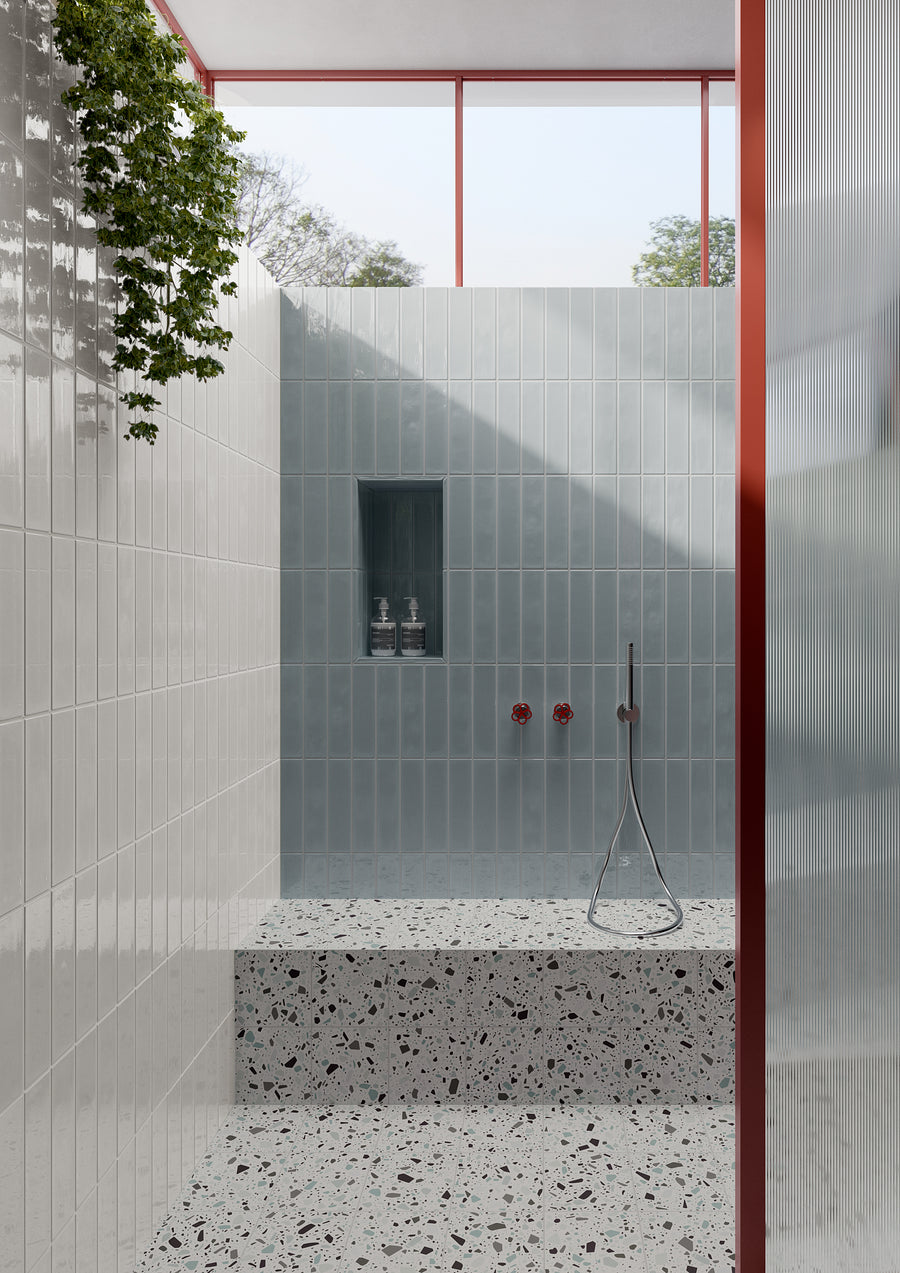 Minimalist shower with light gray vertical tiles, terrazzo steps, built-in shelf with bottles, a modern handheld shower, red fixtures, glass door, and a wall with cascading green plants. Natural light streams from a window above.