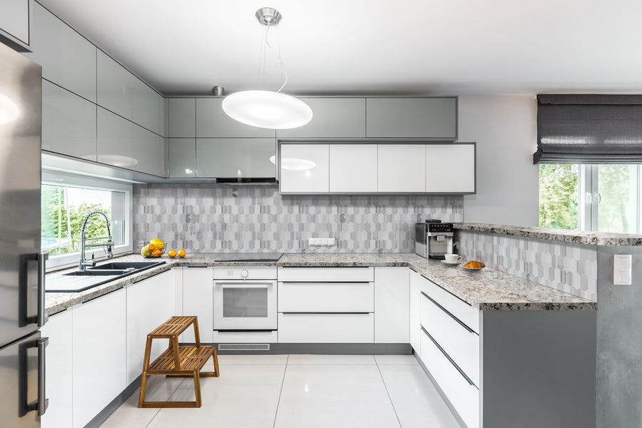 Modern kitchen with white and gray cabinets, geometric tile backsplash, granite countertops, built-in oven, sink, wooden step stools, and a window with a view of greenery. A bowl of oranges sits on the counter.