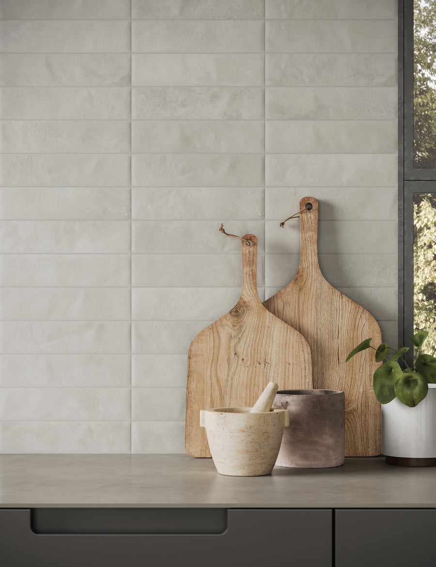 Two wooden cutting boards, a stone mortar and pestle, and a small plant sit on a grey kitchen countertop in front of a white textured tile backsplash and a window.