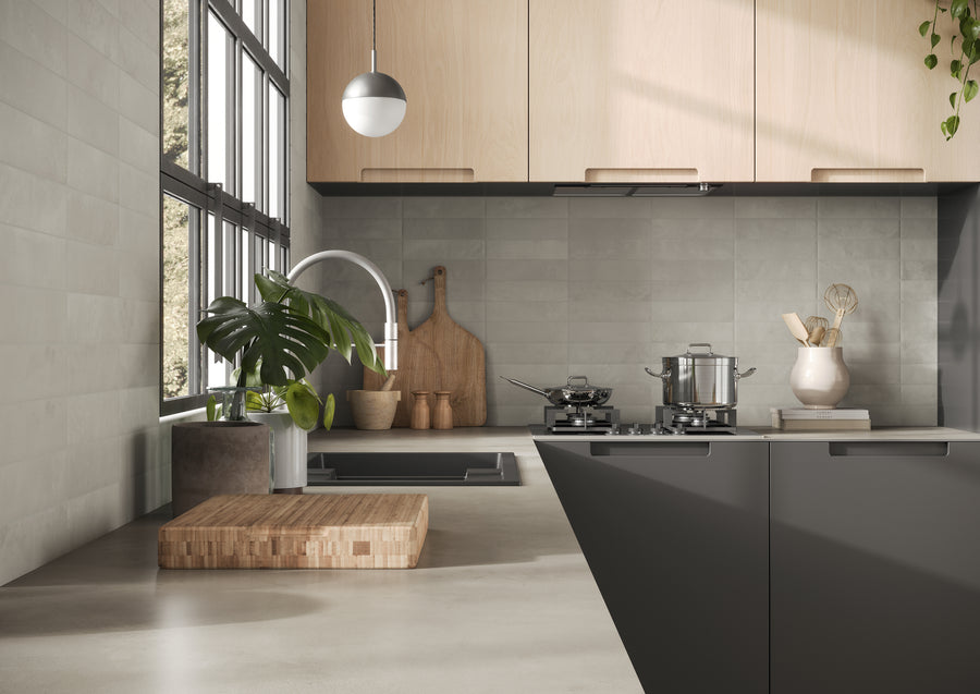 Modern kitchen with a sleek grey countertop, a sink with a silver faucet, a wooden cutting board, potted plant, stovetop with cookware, and light wood cabinets against tiled walls, lit by large windows.