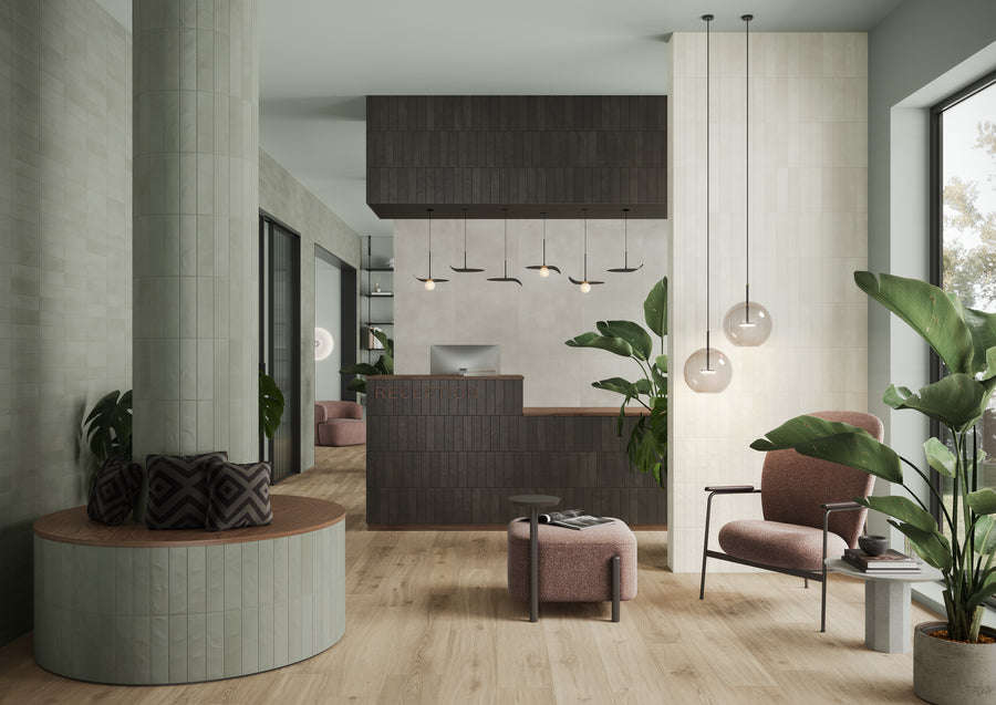 Modern lobby with a wooden reception desk, pendant lights, pink armchairs, large plants, a round bench with cushions, and floor-to-ceiling windows letting in natural light. Light wood flooring and muted green and gray tones throughout.