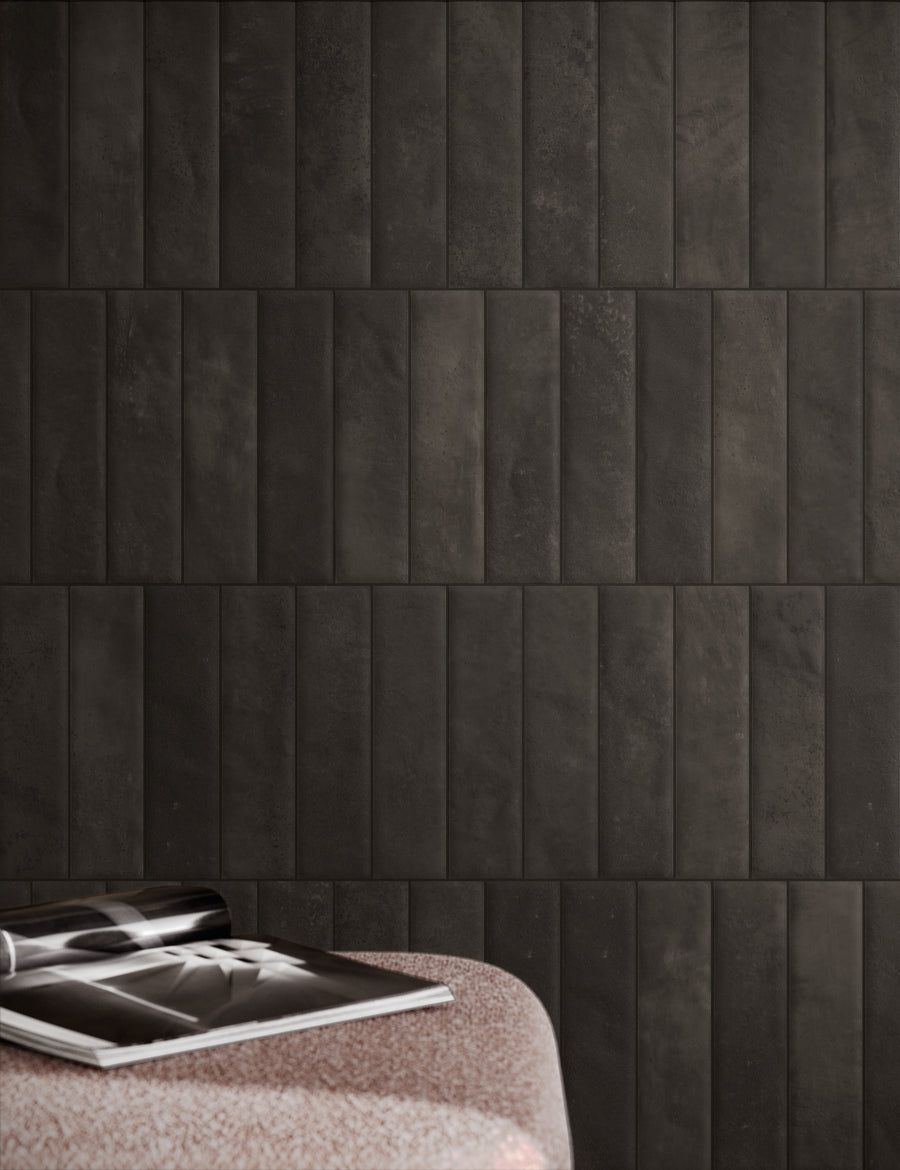 A magazine rests open on a speckled pink surface in front of a wall covered with rectangular, vertically arranged dark gray tiles.