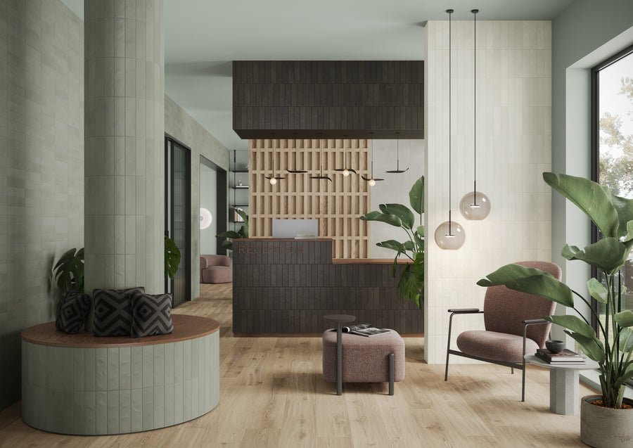 A modern reception area with a wooden desk, a gridded wood accent wall, green plants, a round bench with cushions, two pink armchairs, pendant lights, large windows, and light wood flooring.