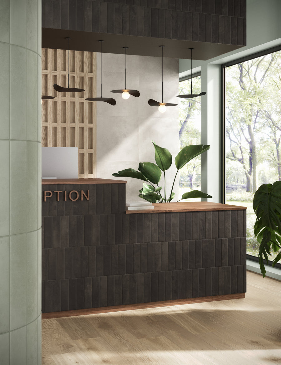 A modern reception desk with dark wood paneling, green plants, pendant lights, and large windows letting in natural light with trees visible outside. The word RECEPTION is partially visible on the desk.