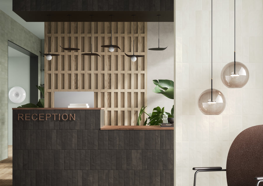Modern reception area with a dark tiled desk, wooden lattice wall, hanging globe lights, large green plants, and a neutral color palette. The word RECEPTION is displayed on the desk front.