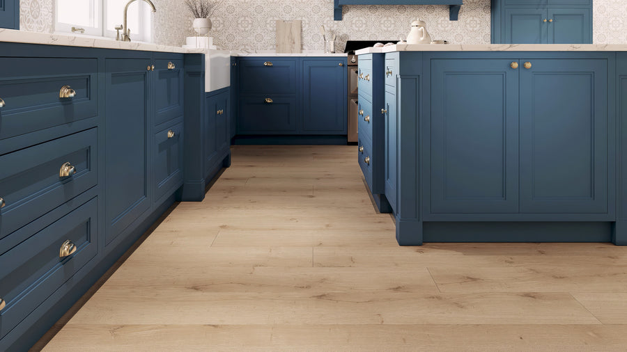 A modern kitchen with blue cabinets, brass handles, white marble countertops, light wood flooring, a farmhouse sink, and patterned wallpaper. The space is bright and tidy.