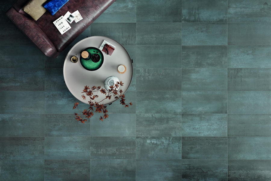 A round coffee table with a vase, small plant, beverages, and snacks sits beside a dark sofa with books and fabric swatches, all on a teal-gray tiled floor viewed from above.