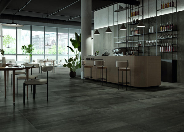 A modern, spacious cafe with large windows, minimalist furniture, a sleek counter with bar stools, indoor plants, and shelves stocked with bottles and glassware, all in neutral and dark tones.