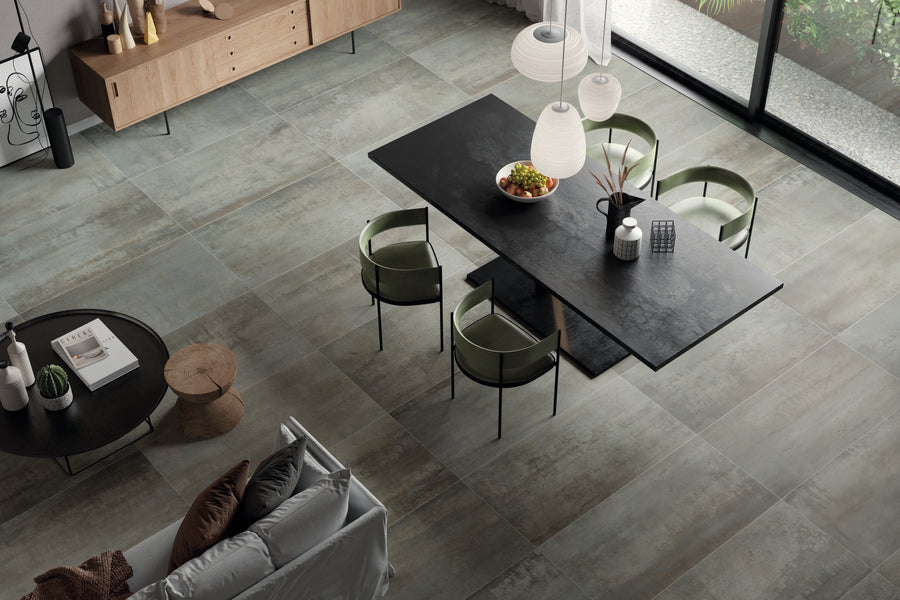 Modern dining area with a black rectangular table, green chairs, pendant lights overhead, and a bowl of fruit. Large textured floor tiles, a beige sofa, and wooden sideboard complete the minimalist, stylish room.