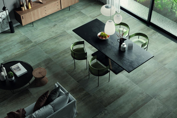 Modern dining room with a black rectangular table, four green chairs, pendant lights, a bowl of fruit, and large floor tiles. Floor-to-ceiling windows let in natural light; minimalist decor and a sideboard are visible.