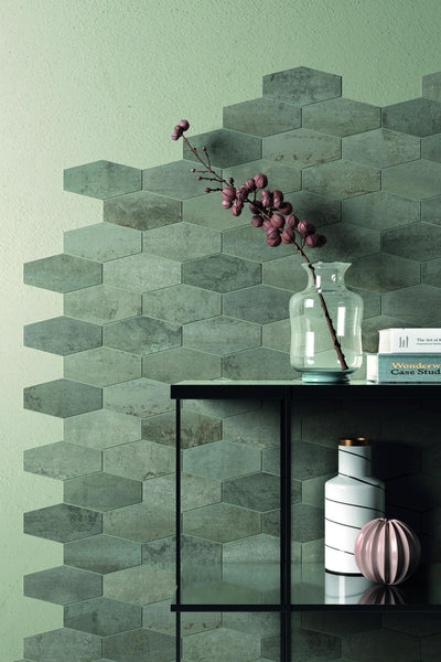 A modern interior with hexagonal green tiles on the wall, a glass vase with a branch of pink flowers, books, and decorative objects on a black metal shelf. The wall is painted a soft green color.