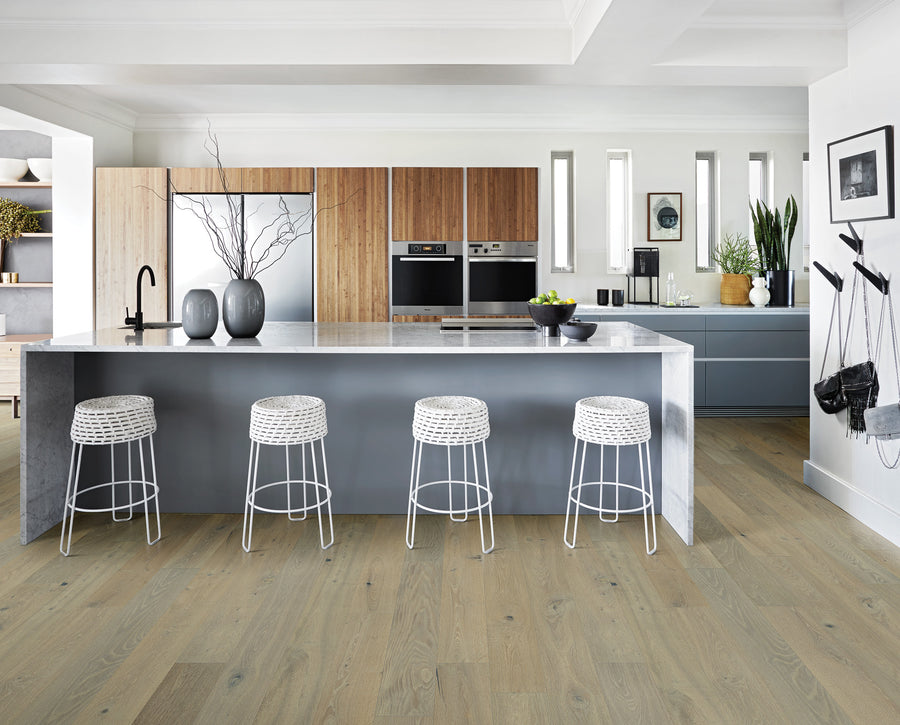 Modern kitchen with a large island, four white woven barstools, wood and gray cabinets, built-in appliances, plants, and decorative vases, all set on light wood flooring.