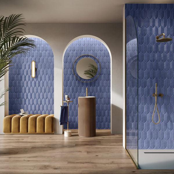 A modern bathroom with blue scalloped tile walls, arched alcoves, a round mirror above a cylindrical sink, a gold shower, a gold bench, and wooden flooring. A palm plant adds greenery to the stylish space.