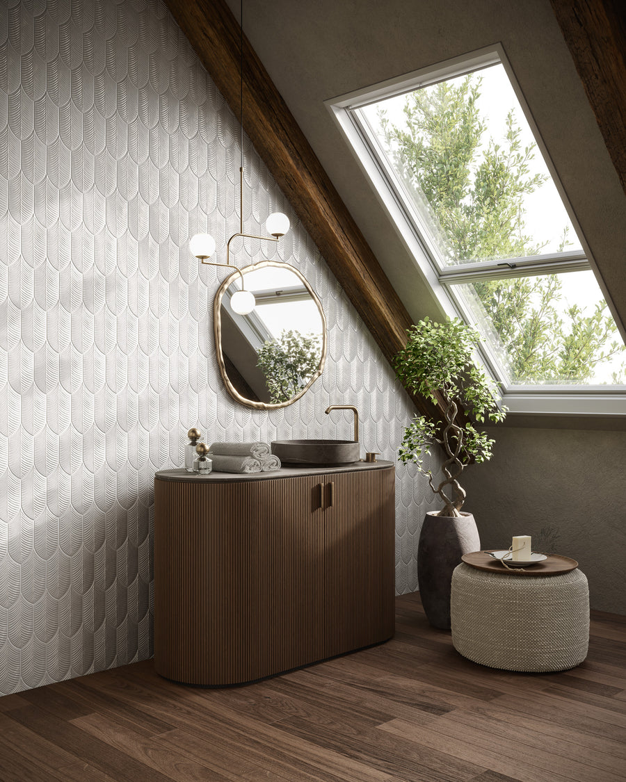 A modern attic bathroom with a sloped ceiling, skylight window, round mirror, pendant light, wood vanity with sink, textured white tile wall, potted plant, and woven stool with towel and soap.
