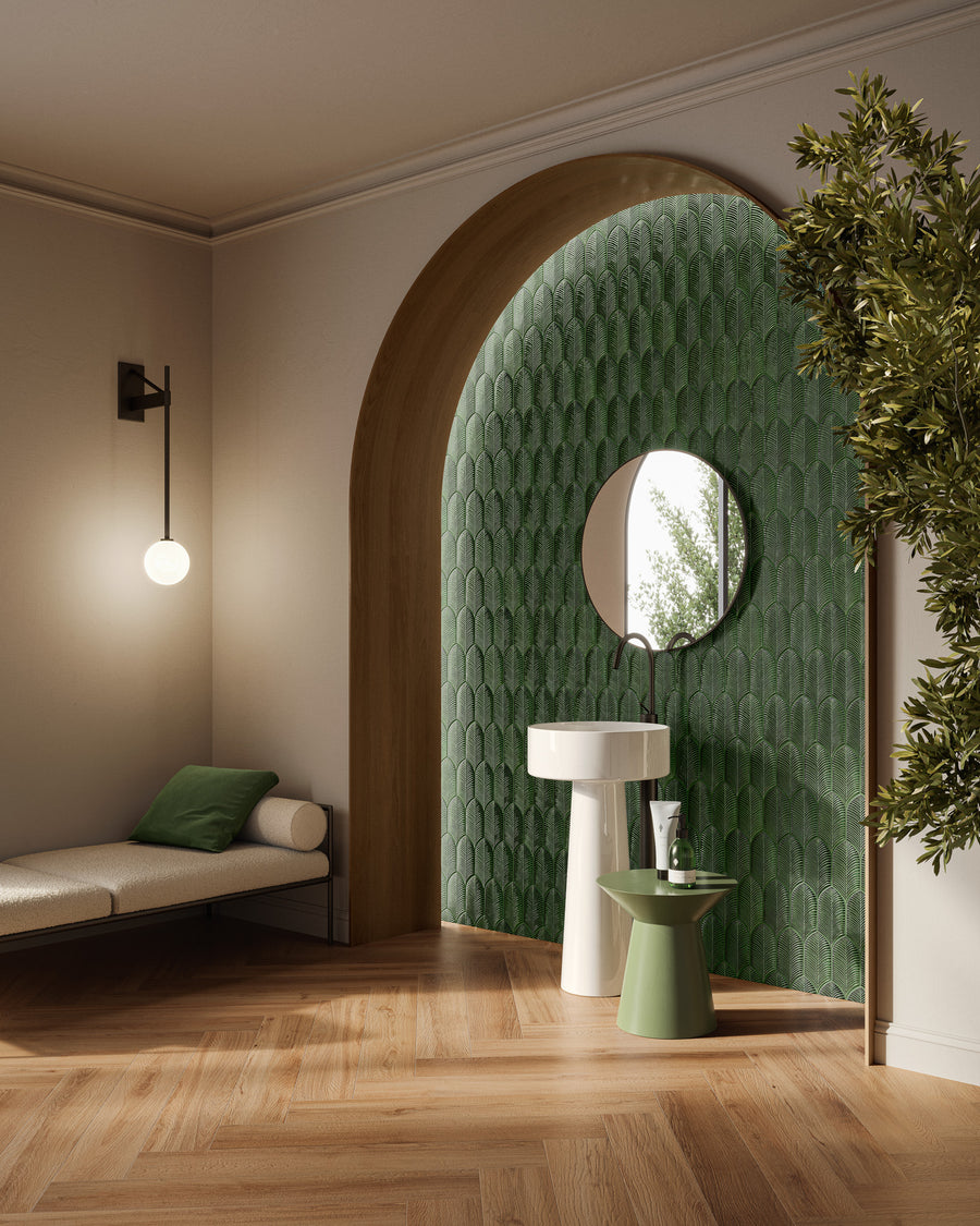 A modern bathroom with a green tiled arched accent wall, round mirror, white pedestal sink, and a small green side table. A bench with green pillows and a wall lamp are on the left; a leafy plant is on the right.