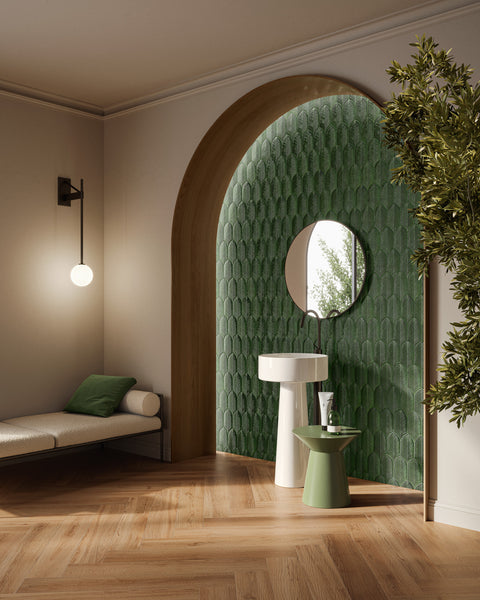 A modern bathroom with a green tiled arched accent wall, round mirror, white pedestal sink, and a small green side table. A bench with green pillows and a wall lamp are on the left; a leafy plant is on the right.