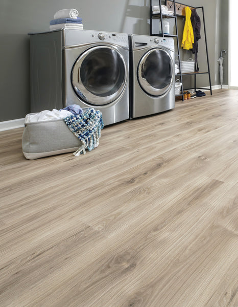 A modern laundry room with a front-loading washer and dryer, a basket with towels and a blanket on the floor, and shelves holding laundry supplies and clothes against a gray wall. The floor is light wood.