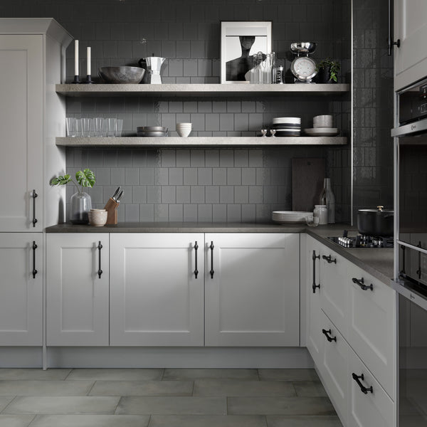Modern kitchen with gray tiled backsplash, white cabinets, and black handles. Two open shelves hold dishes, glasses, kitchenware, and decorative items. Countertop has a plant, cutting board, and kitchen scale.