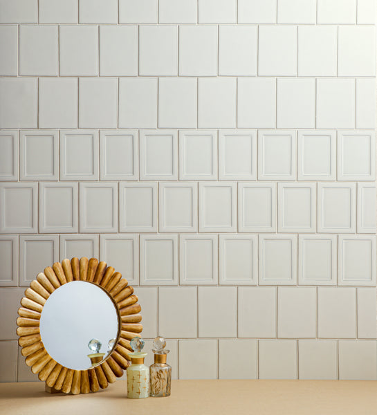 A round mirror with a gold, petal-like frame sits on a beige surface next to three vintage glass perfume bottles, with a white tiled wall in the background.