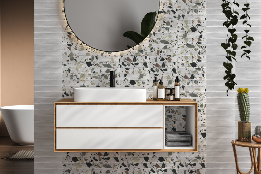 Modern bathroom with a round mirror, terrazzo tile backsplash, white sink on a wooden vanity with toiletries, folded towels, and plants as decor. White tub and cactus visible in the background.