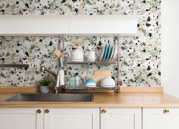 A modern kitchen with white cabinets, a wood countertop, a stainless steel sink, and a dish rack holding plates, bowls, cups, and kitchen utensils in front of a terrazzo-patterned backsplash.