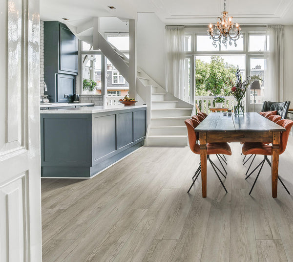 Bright, modern kitchen and dining area with large windows, light wood flooring, a gray island, white staircase, wooden dining table, orange chairs, and a chandelier. Natural light fills the open, airy space.