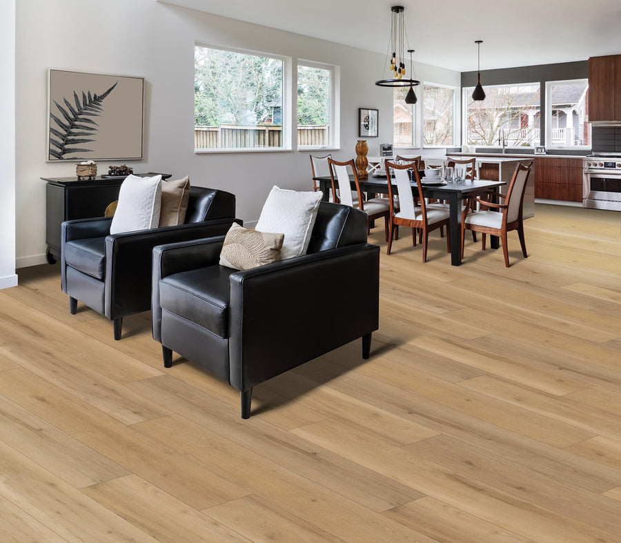 Modern open-concept living and dining area with light wood flooring, two black armchairs with pillows, a dining table with eight chairs, large windows, and a kitchen with wood cabinets and stainless steel appliances.