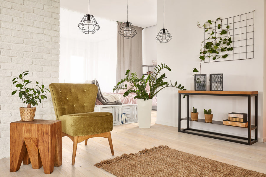 A cozy, modern living room with an olive green armchair, wooden side table, potted plants, a jute rug, and a console table with decor. Three pendant lights and a grid wall frame with vines complete the airy space.