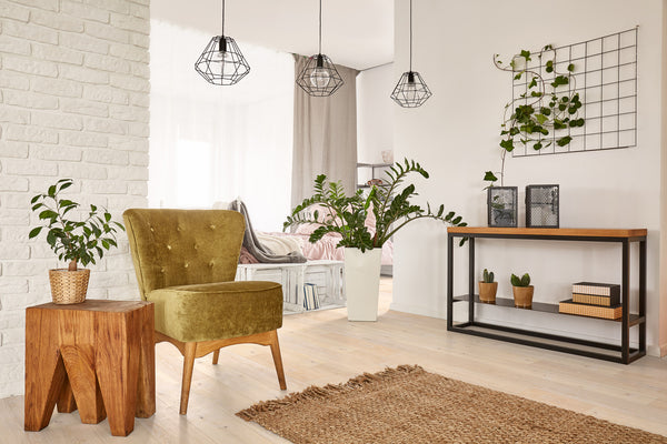 A cozy, modern living room with an olive green armchair, wooden side table, potted plants, a jute rug, and a console table with decor. Three pendant lights and a grid wall frame with vines complete the airy space.