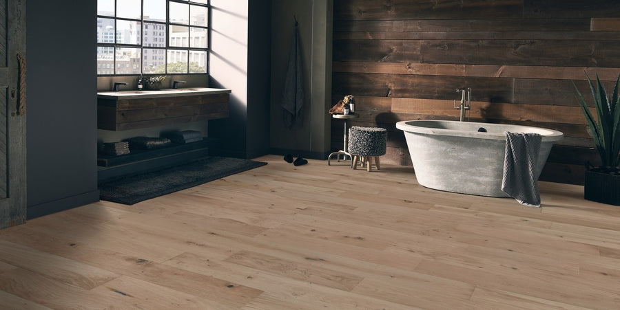 A modern bathroom with wide light wood floors, a rustic wood-paneled wall, a freestanding gray bathtub, a wooden vanity with a sink, a large window, and a potted plant in the corner.
