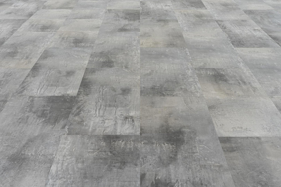 A close-up of a floor showcases Stonescape Luxury Vinyl Plank, featuring rectangular gray tiles in a staggered pattern with a textured, weathered look that captures the appeal of durable stone-inspired flooring.