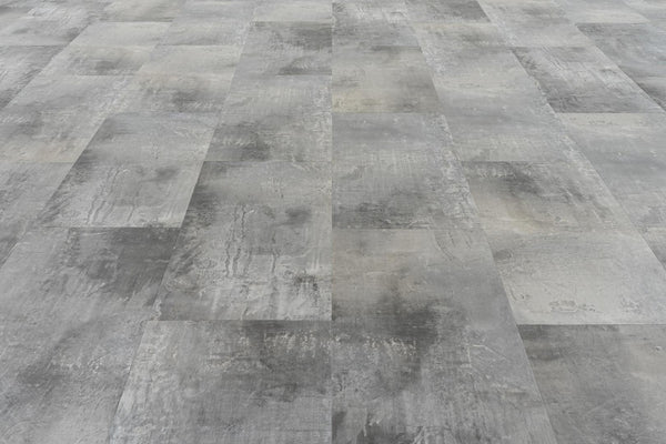 A close-up of a floor showcases Stonescape Luxury Vinyl Plank, featuring rectangular gray tiles in a staggered pattern with a textured, weathered look that captures the appeal of durable stone-inspired flooring.