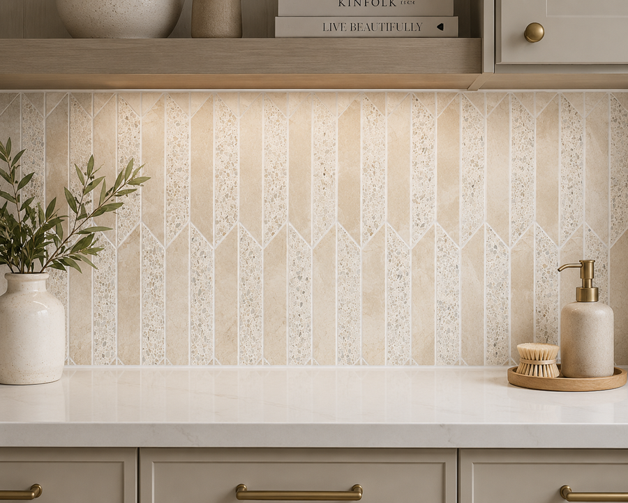 Beige geometric tile backsplash featuring the Revive Terrazzo & Korbell Half Picket Mosaic, complemented by a white countertop with a soap dispenser, brush, and potted plant. The upper shelf displays books, a bowl, and a vase, while warm under-cabinet lighting highlights the clean, modern kitchen decor.