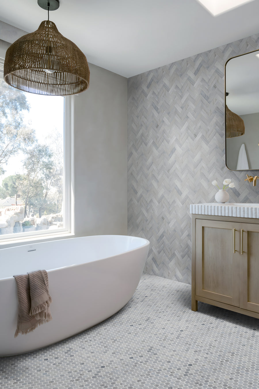 Modern bathroom with a freestanding white tub, taupe towel, large window, rattan pendant light, wood vanity with striped countertop, and a Blue Horizon 1" Pennyround Honed Marble Tile accent wall for added texture.