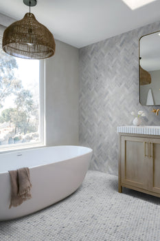 Modern bathroom with a freestanding white tub, taupe towel, large window, rattan pendant light, wood vanity with striped countertop, and a Blue Horizon 1