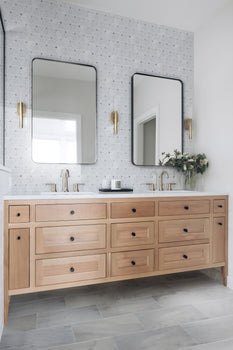 Modern bathroom with a light wood double-sink vanity, two black-framed rectangular mirrors, brass wall sconces, a Blue Horizon 12x24 Honed Marble Tile backsplash in blue, and a vase of flowers on the counter.