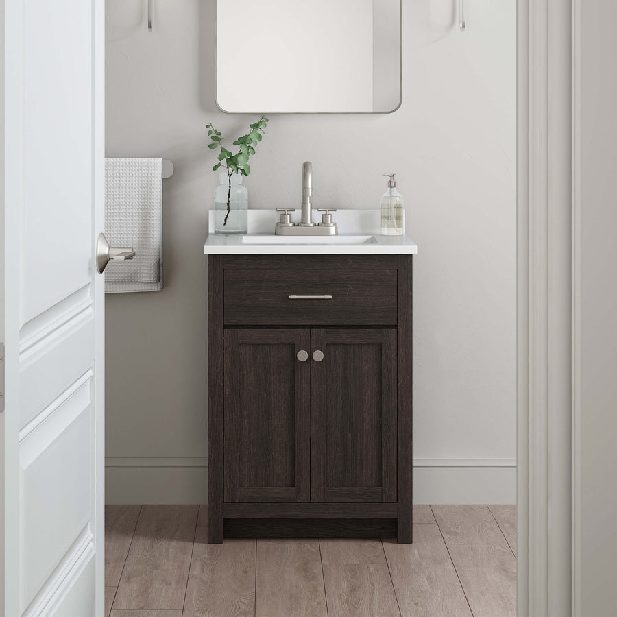 A small bathroom features a dark wood vanity with a white sink, silver faucet, soap dispenser, and a leafy plant. A square mirror hangs above, and a towel is folded on a rail to the left. The floor is light wood.