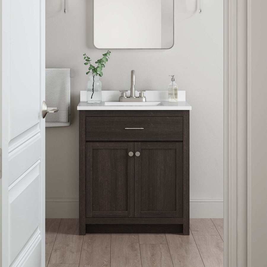 A small bathroom vanity with a dark wood cabinet, white countertop, silver faucet, and a rectangular mirror above. A towel hangs on the wall, and a plant and soap dispenser are on the sink. Light wood floor.
