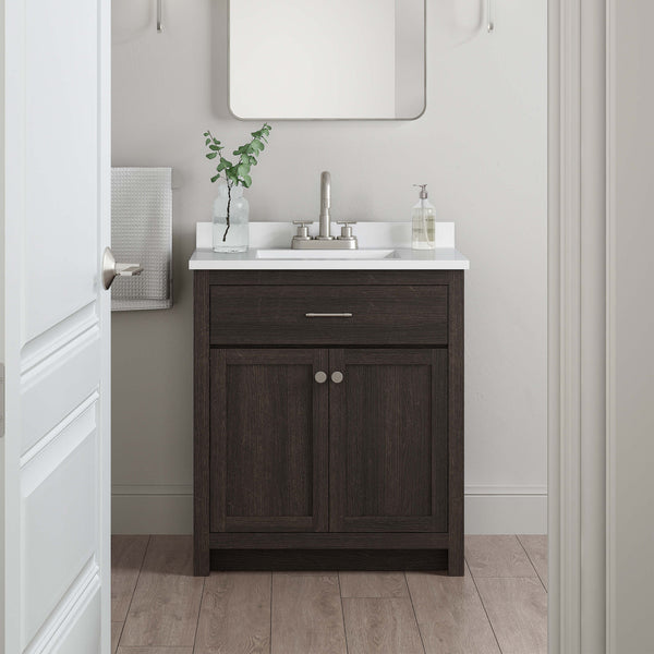 A small bathroom vanity with a dark wood cabinet, white countertop, silver faucet, and a rectangular mirror above. A towel hangs on the wall, and a plant and soap dispenser are on the sink. Light wood floor.