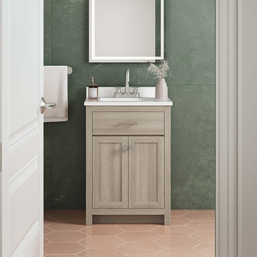 A small bathroom with green marble walls and peach hexagon tile floor. Theres a light wood vanity with a white countertop, a mirror above, a faucet, soap dispenser, and a vase with dried flowers. A towel hangs on the left wall.