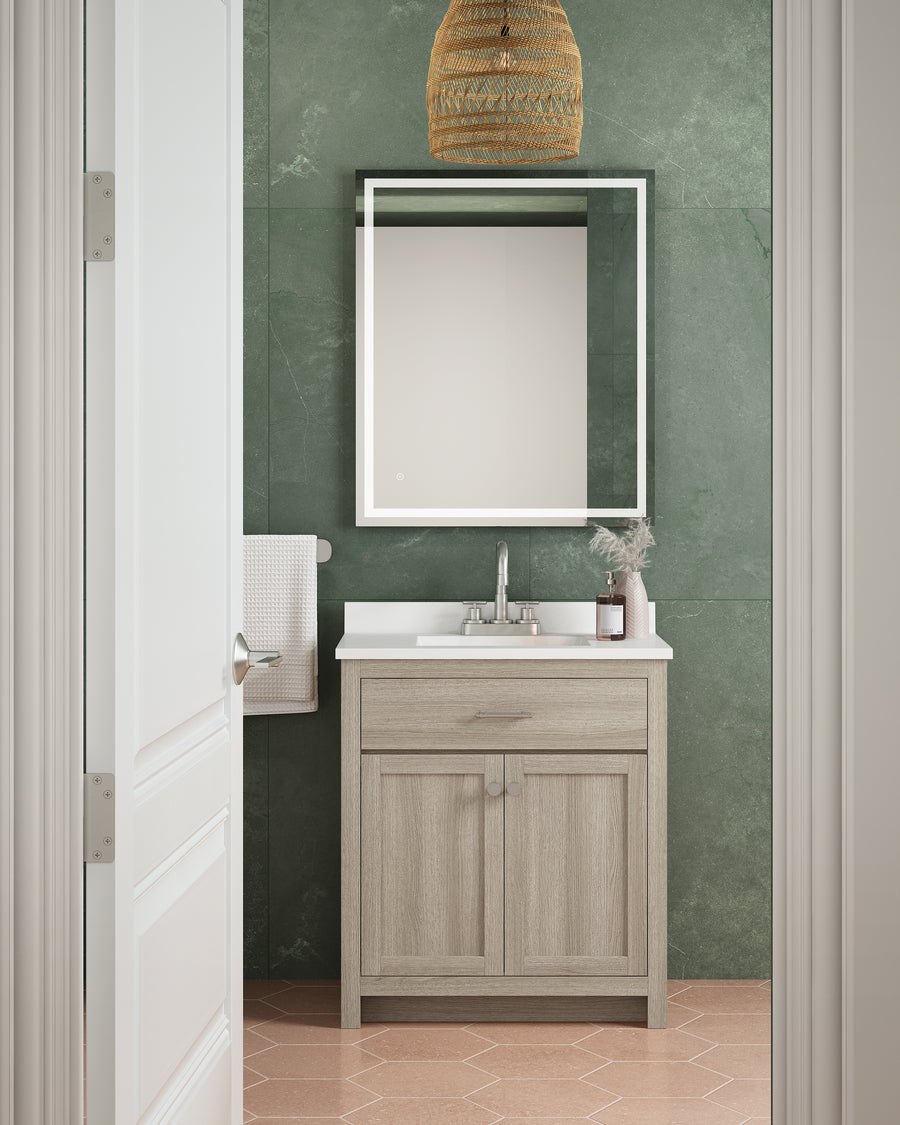 A small green-walled bathroom showcases a Reese 30" Vanity with a white sink, rectangular mirror, towel rack, pendant light, and a plant on the counter, all visible through an open white door.