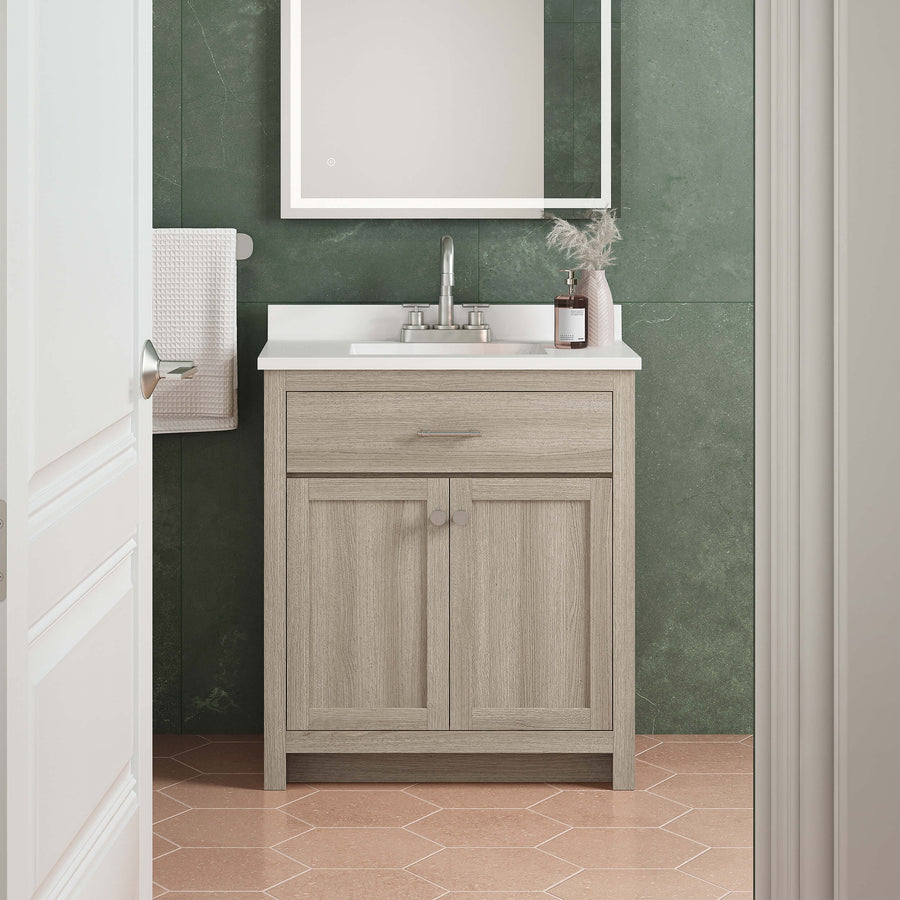 A small bathroom vanity with a light wood cabinet, white countertop, and chrome faucet sits against a green tiled wall. A framed mirror hangs above, and a towel is neatly folded on a wall rack. The floor has peach hexagon tiles.