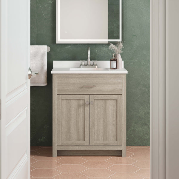 A small bathroom vanity with a light wood cabinet, white countertop, and chrome faucet sits against a green tiled wall. A framed mirror hangs above, and a towel is neatly folded on a wall rack. The floor has peach hexagon tiles.