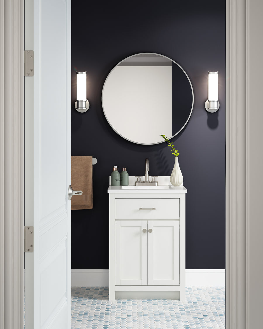 A modern bathroom features dark navy walls, a round mirror, two wall sconces, and the space-saving Reese 24" Vanity with sink. Greenery in a vase, toiletries, a brown towel, and patterned blue-and-white tile flooring complete the look.