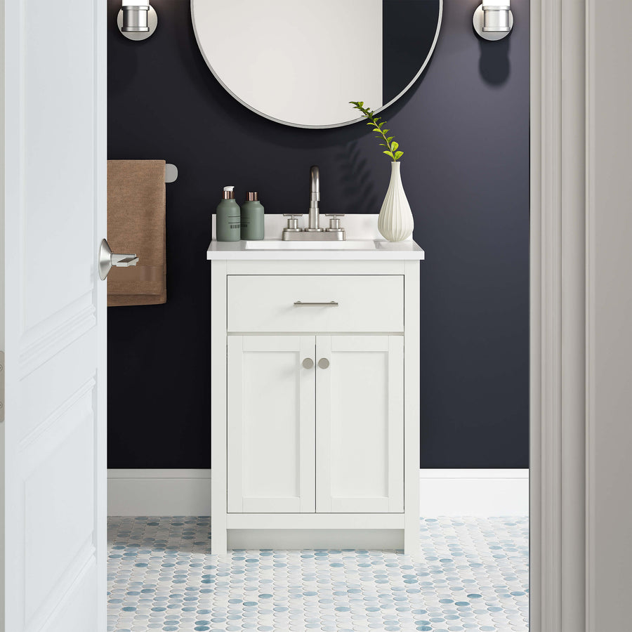 A small bathroom with a white vanity, silver faucet, round mirror, dark wall, and hexagon-patterned tile floor. A vase with a green branch and toiletries sit on the vanity; a brown towel hangs on the wall.
