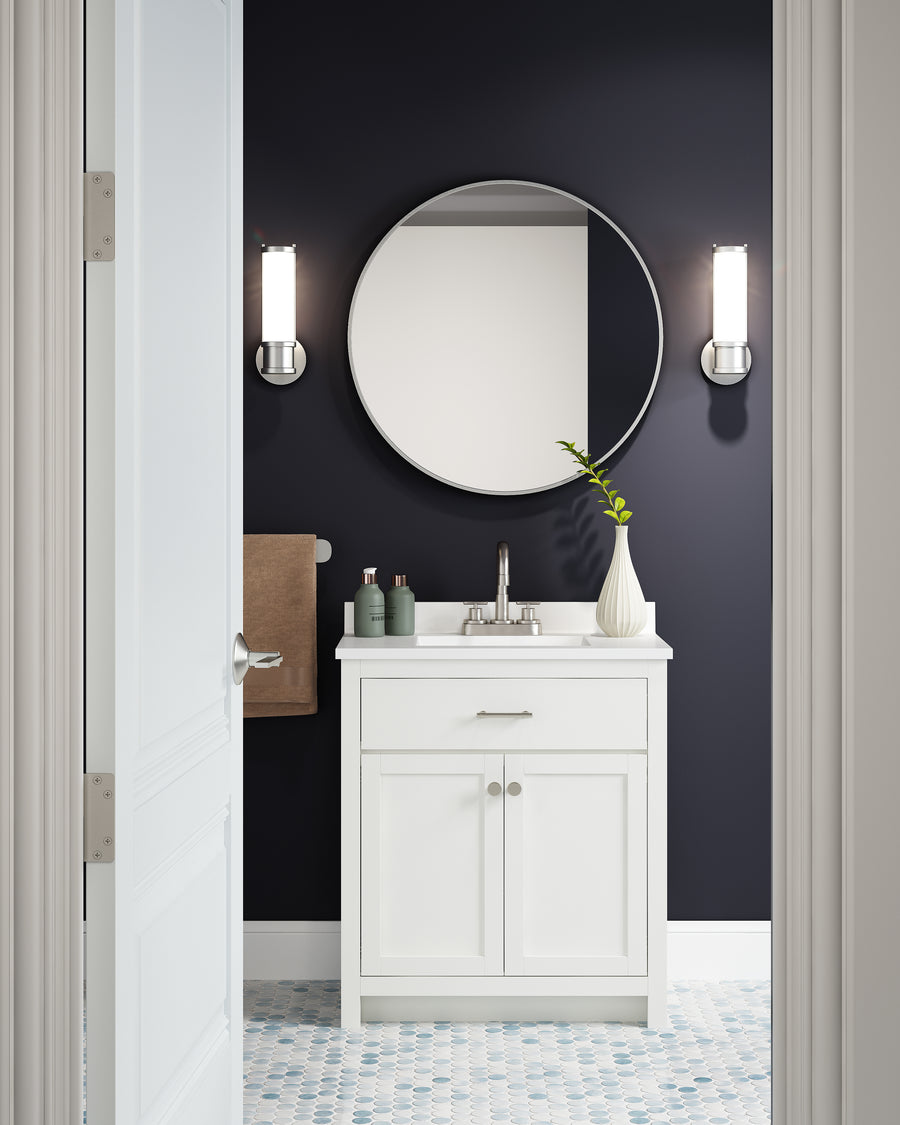 A modern bathroom featuring the Reese 30" Vanity with a white finish, a round mirror, two wall sconces, a brown towel, and a vase with a green stem—all set against a dark navy wall and patterned tile floor.